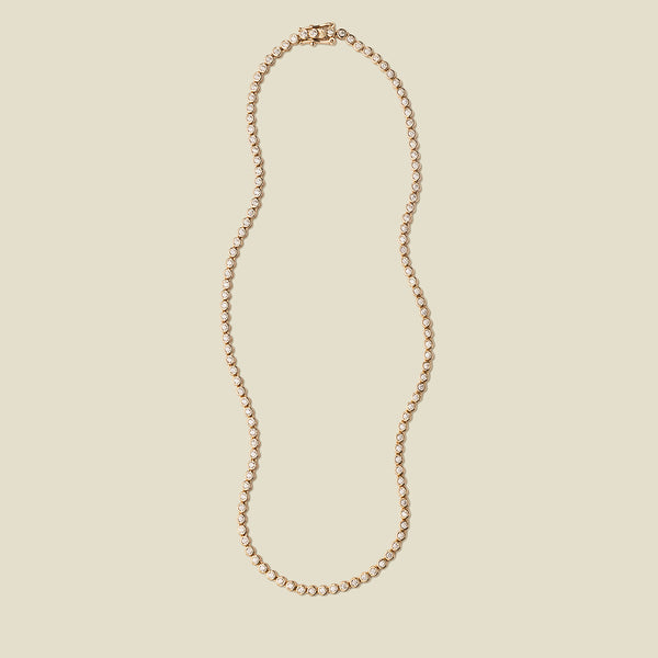 A gold tennis necklace with round bezel-set clear gemstones, set against a solid beige background.