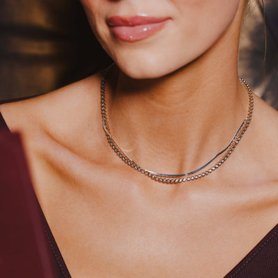 A close-up of a person in a burgundy top wearing a wide flat gold necklace layered with a gold tennis necklace with round bezel-set clear gemstones.