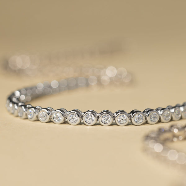 A close-up of a silver tennis necklace, showcasing the round bezel-set clear gemstones, laying on a solid beige surface.