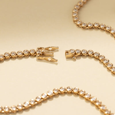 A gold tennis necklace laying on a solid beige background, showing the box clasp open.