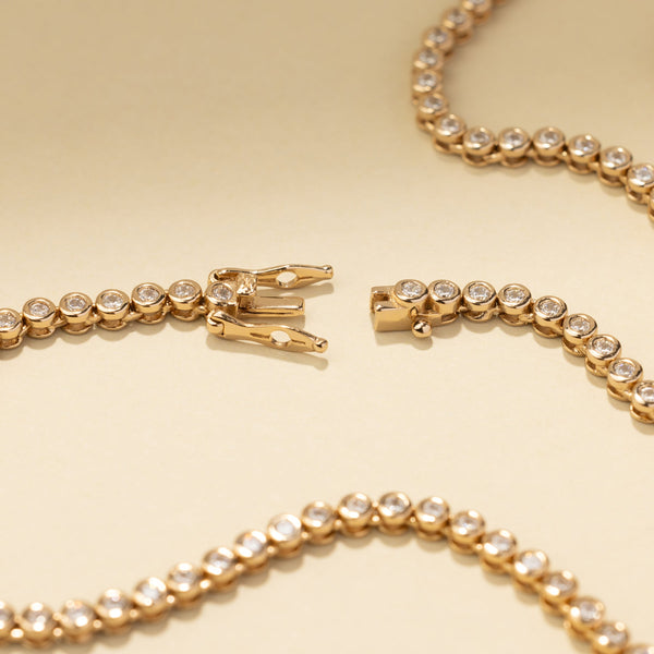 A gold tennis necklace laying on a solid beige background, showing the box clasp open.