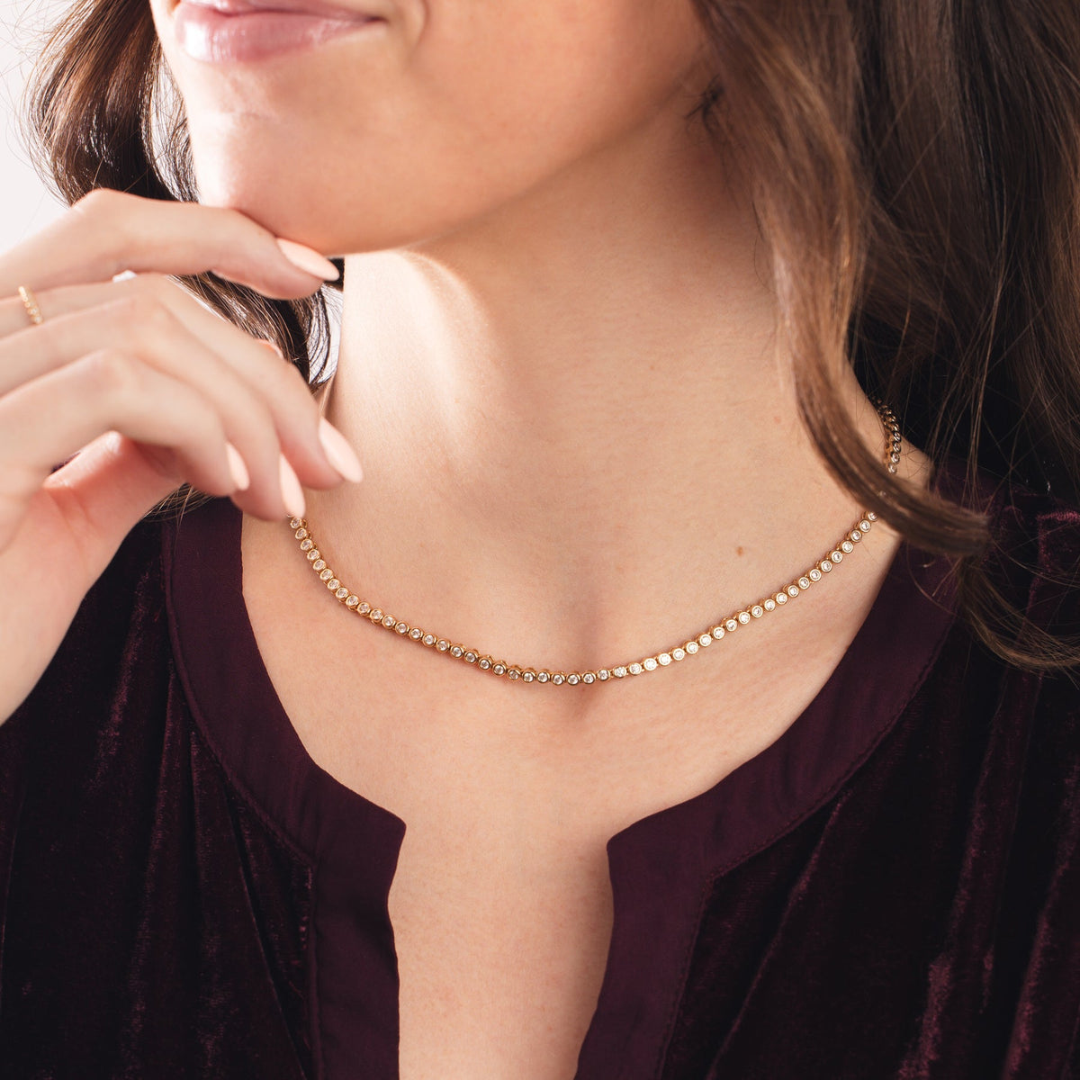A close-up of a person in a burgundy shirt wearing a gold tennis necklace with round bezel-set clear gemstones.