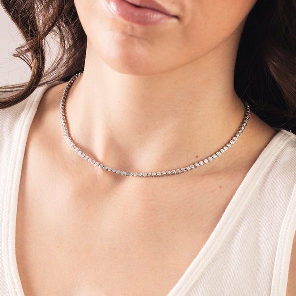 A close-up of a person wearing a silver tennis necklace with round bezel-set clear gemstones 