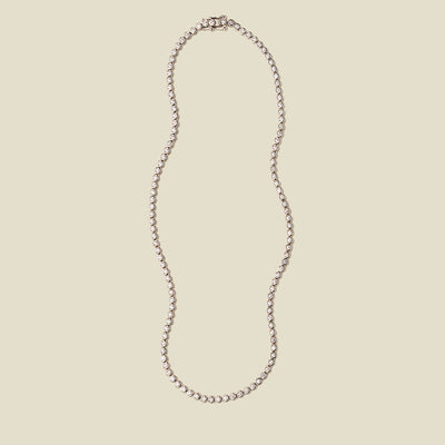 A silver tennis necklace with round bezel-set clear gemstones, set against a solid beige background.