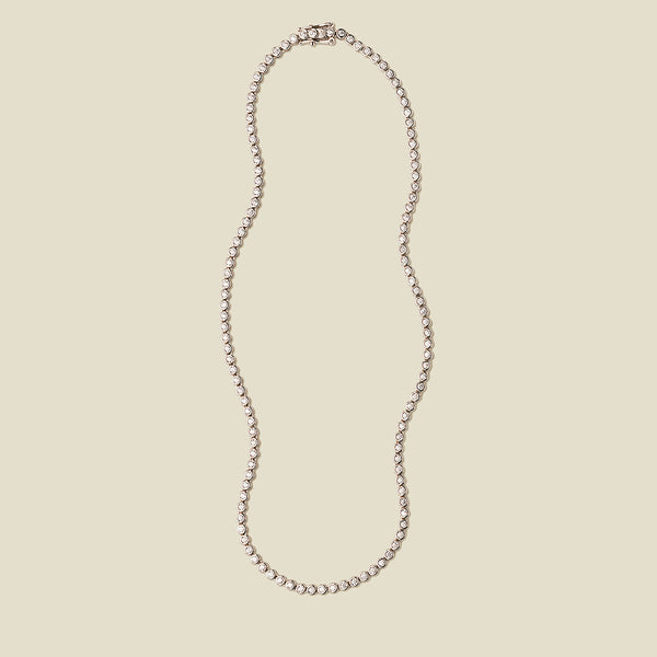 A silver tennis necklace with round bezel-set clear gemstones, set against a solid beige background.