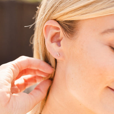 A close-up of a person's ear wearing a gold stud with three clear baguette-cut gemstones.