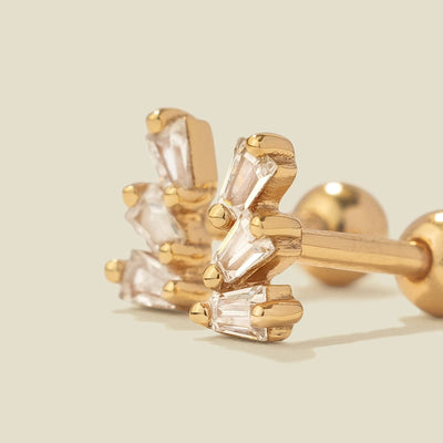 A pair of gold-colored stud earrings with baguette-cut clear stones, shown on a solid light beige background.