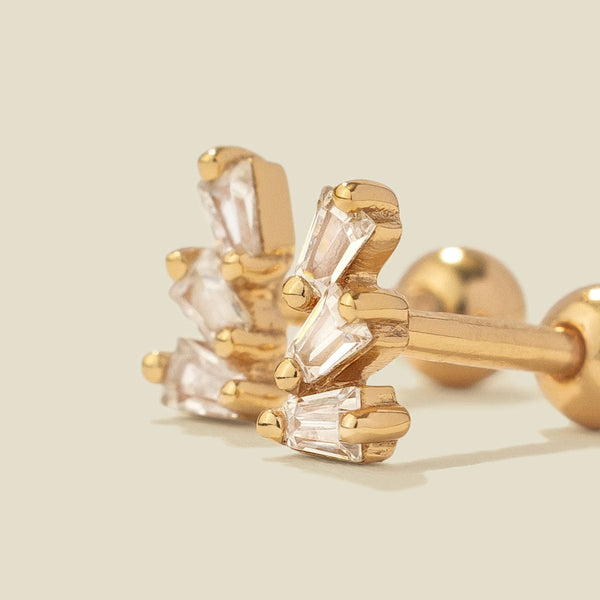 A pair of gold-colored stud earrings with baguette-cut clear stones, shown on a solid light beige background.