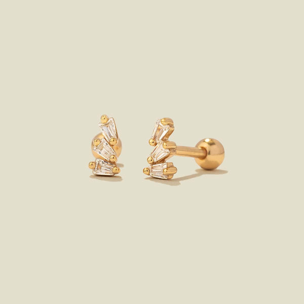 A pair of gold-colored stud earrings with baguette-cut clear stones, shown on a solid light beige background.