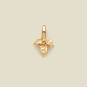 A polished gold holly leaf pendant charm with a hanging loop, centered on a solid light beige background.