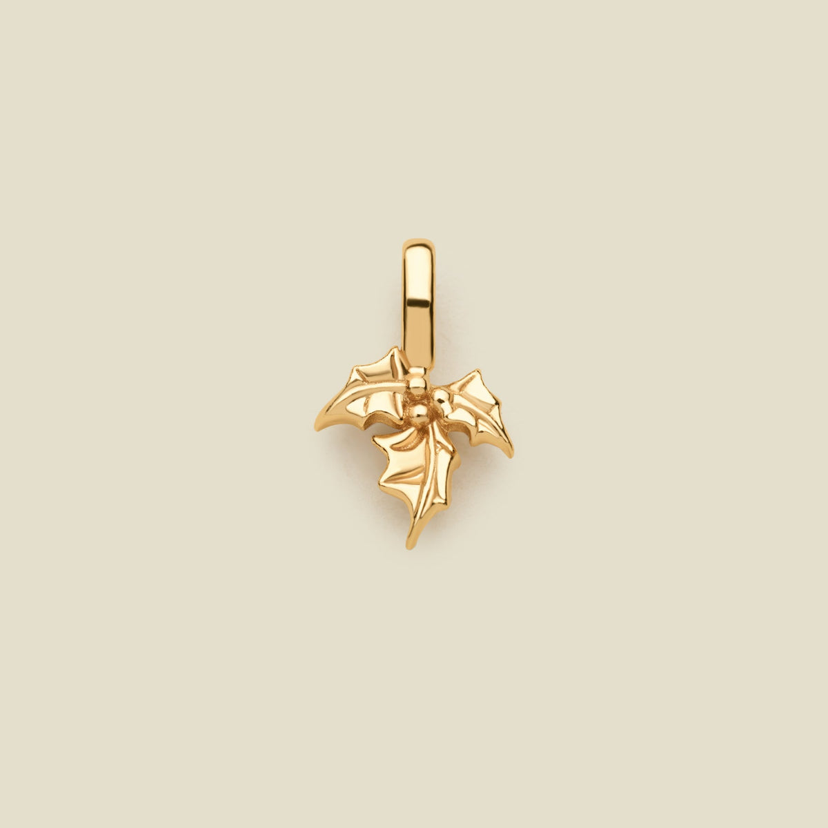 A polished gold holly leaf pendant charm with a hanging loop, centered on a solid light beige background.