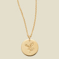 A gold chain necklace with a round gold disc pendant engraved with a holly leaf design, shown against a solid light beige background.