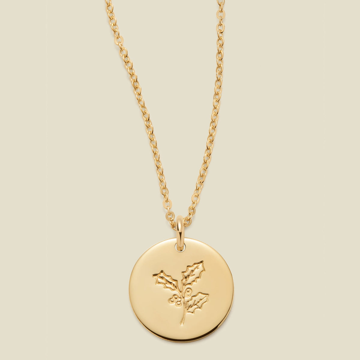 A gold chain necklace with a round gold disc pendant engraved with a holly leaf design, shown against a solid light beige background.