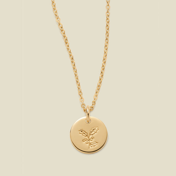 A gold chain necklace with a round gold disc pendant engraved with a holly leaf design, shown against a solid light beige background.