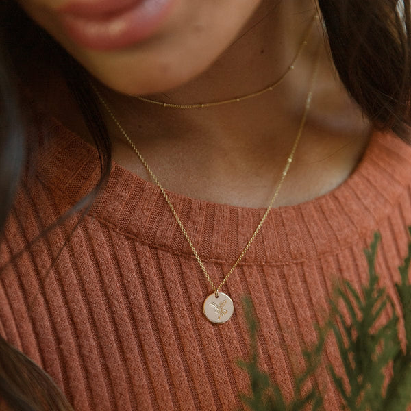 A close-up of a woman wearing a burnt orange ribbed top with a dainty gold choker necklace and a longer pendant chain featuring a round flat gold disc. The disc is engraved with a holly floral design.