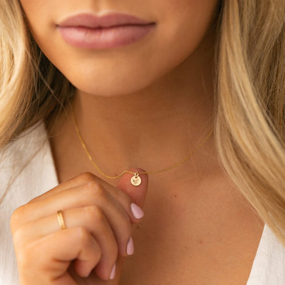 A close-up of a blonde woman holding the small round flat engraved disc on the gold pendant chain she is wearing. The disc has a holly floral motif.