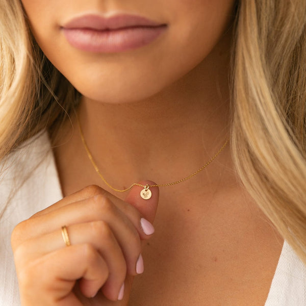 A close-up of a blonde woman holding the small round flat engraved disc on the gold pendant chain she is wearing. The disc has a holly floral motif.