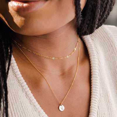 A close-up of a woman in a cream knit sweater wearing a textured gold choker necklace layered with a gold pendant chain featuring a round flat disc charm. The disc is engraved with a holly leaf design.