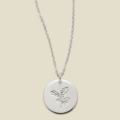 A silver chain necklace with a round silver disc pendant engraved with a holly leaf design, shown against a solid light beige background.