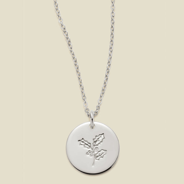 A silver chain necklace with a round silver disc pendant engraved with a holly leaf design, shown against a solid light beige background.
