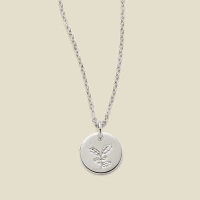 A silver chain necklace with a round silver disc pendant engraved with a holly leaf design, shown against a solid light beige background.