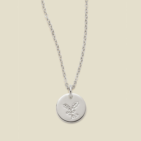 A silver chain necklace with a round silver disc pendant engraved with a holly leaf design, shown against a solid light beige background.