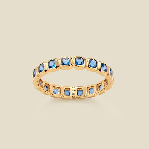 A gold eternity ring featuring a continuous row of square cut blue gemstones, shown on a solid light beige background.