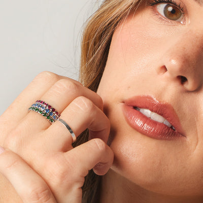 A close-up of a person holding their hand up to their face, showcasing a stack of three silver eternity style rings, each with a different color of square cut gemstones, and single wide silver band ring.