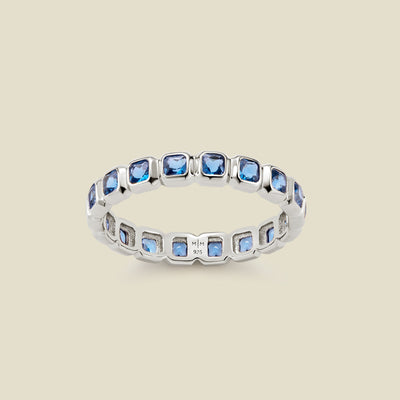 A silver eternity ring featuring a continuous row of square cut blue gemstones, shown on a solid light beige background.