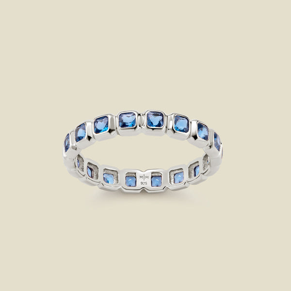 A silver eternity ring featuring a continuous row of square cut blue gemstones, shown on a solid light beige background.