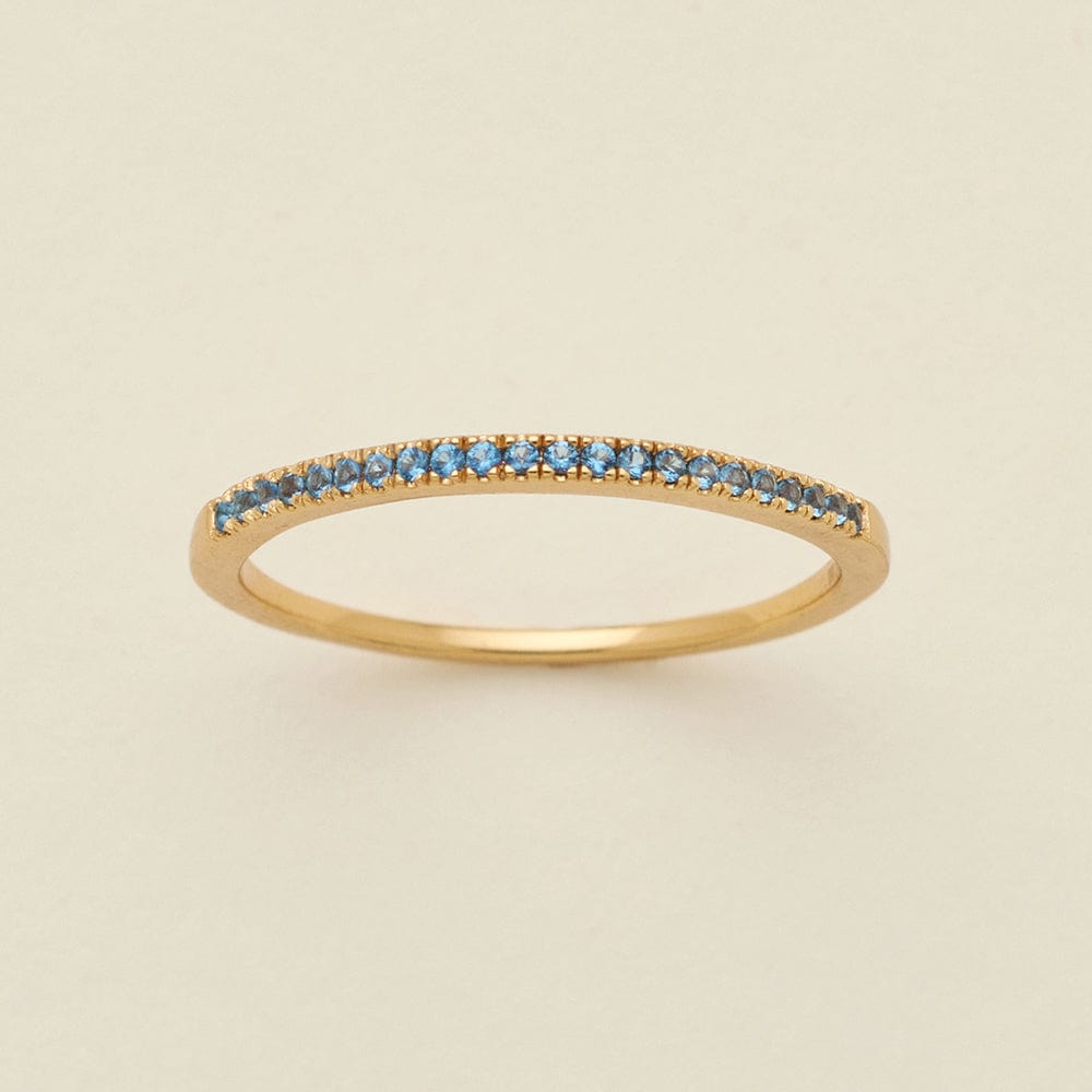 A gold ring featuring a single row of small round-cut light blue gemstones, photographed on a clean, solid, warm off-white background.
