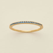 A gold ring featuring a single row of small round-cut light blue gemstones, photographed on a clean, solid, warm off-white background.