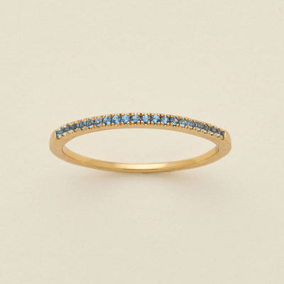 A gold ring featuring a single row of small round-cut light blue gemstones, photographed on a clean, solid, warm off-white background.