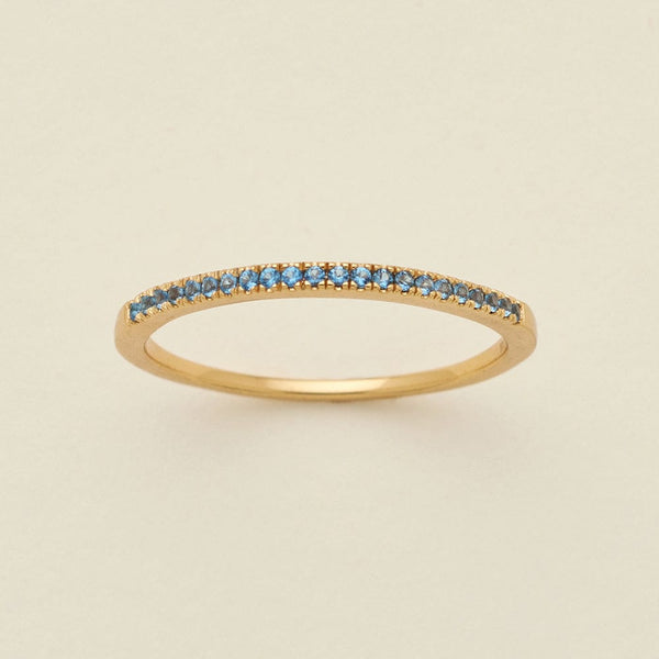 A gold ring featuring a single row of small round-cut light blue gemstones, photographed on a clean, solid, warm off-white background.