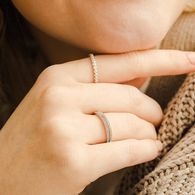 A close-up of a hand resting against a chin, showcasing an eternity band pearl ring and two stacked gold rings, one with small blue gemstones and the other with light green gemstones.