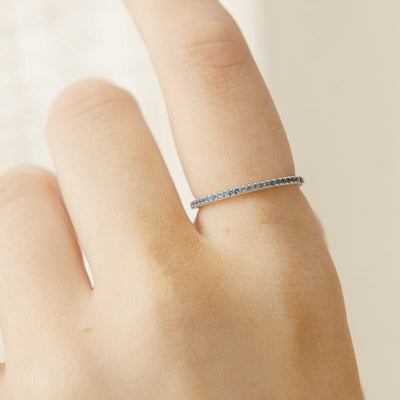 A close-up of a hand wearing a silver ring featuring a single row of small round-cut light blue gemstones, photographed on a clean, white background.