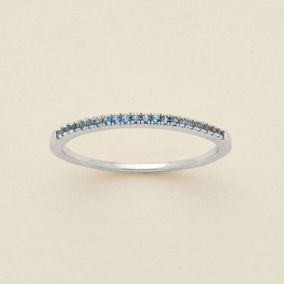 A silver ring featuring a single row of small round-cut light blue gemstones, photographed on a clean, solid, warm off-white background.