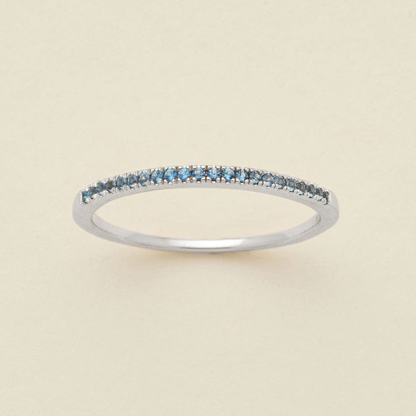 A silver ring featuring a single row of small round-cut light blue gemstones, photographed on a clean, solid, warm off-white background.