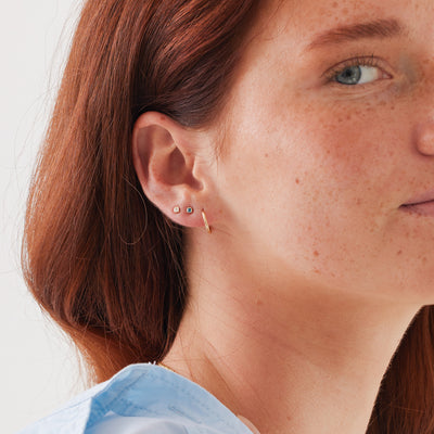 A close-up of a woman's ear wearing a gold hoop earring and two square bezel-set gemstone stud earrings, one in dark blue and one in light blue, set against a solid white background.