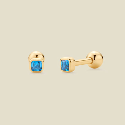A pair of gold stud earrings with square-cut blue gemstones, shown on a light beige background. One earring is facing forward, and the other is turned to the side to show the post and backing.
