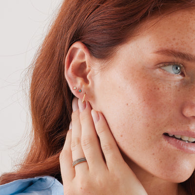 A close-up of a woman's ear wearing a gold paperclip shaped hoop earring and two gold square bezel-set gemstone stud earrings, one with a bright blue stone and the other with light blue.