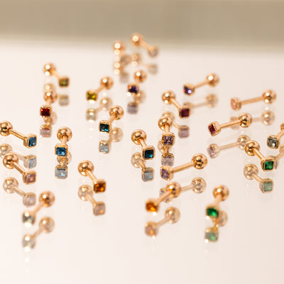 A variety of gold stud earrings, each with a different color of square bezel-set gemstone and a round ball backing, laying on a mirrored surface.