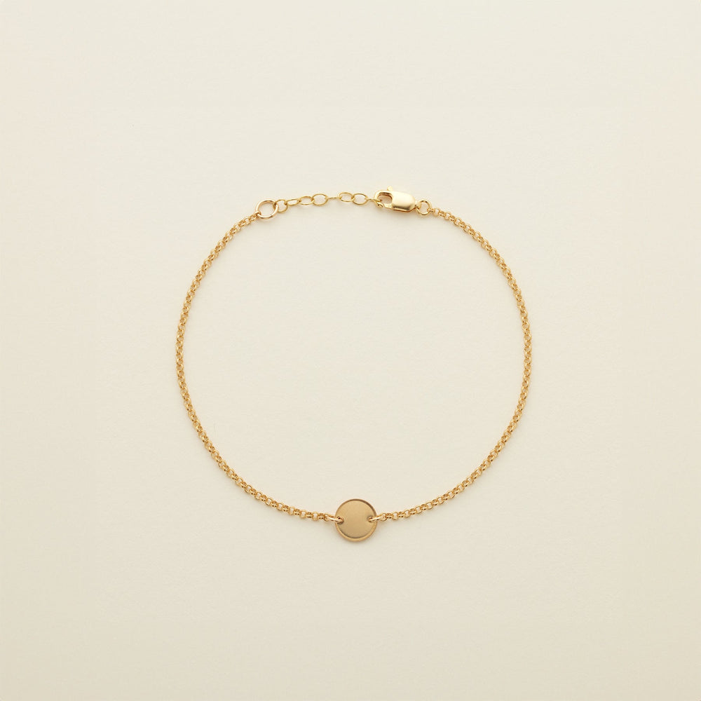 A gold chain anklet with a small round flat disc in the center, shown on a solid light beige background.