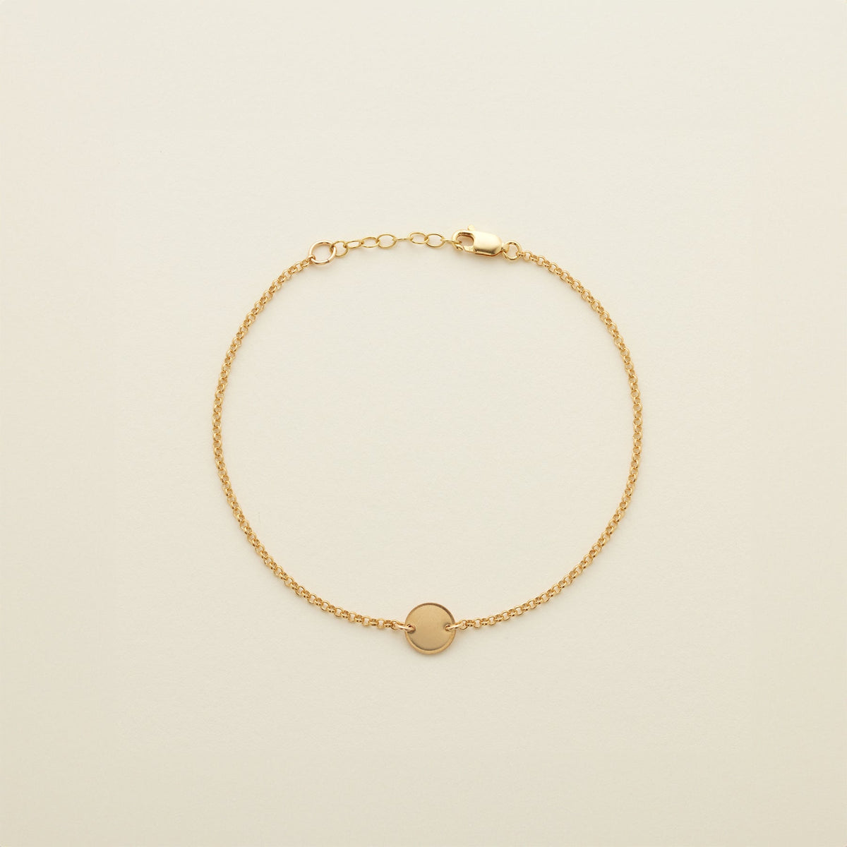 A gold chain anklet with a small round flat disc in the center, shown on a solid light beige background.
