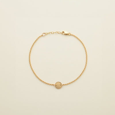 A gold chain anklet with a small round flat disc in the center, shown on a solid light beige background.