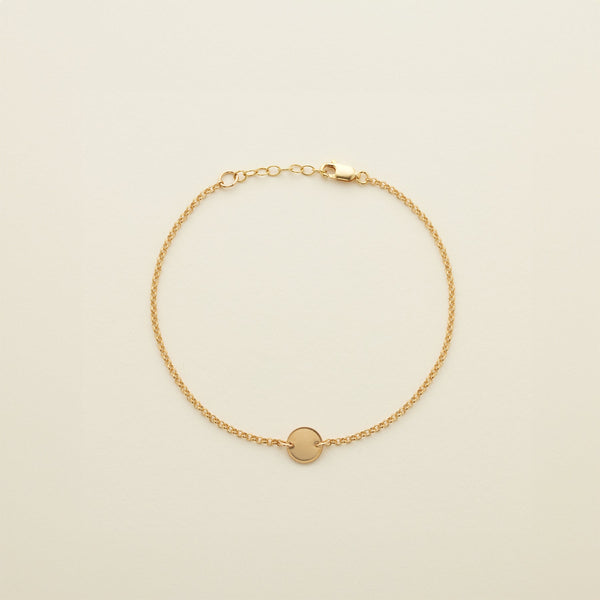 A gold chain anklet with a small round flat disc in the center, shown on a solid light beige background.