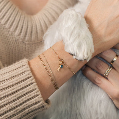 A person's wrist with a white dog paw resting on it, wearing three gold chain bracelets.