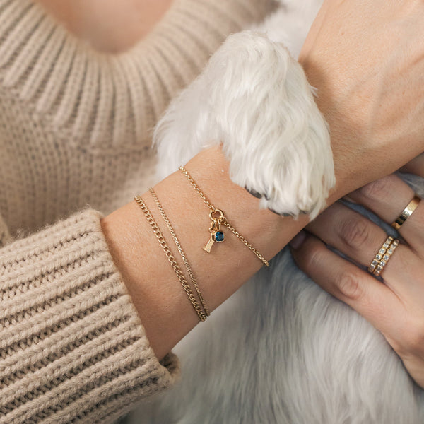 A person's wrist with a white dog paw resting on it, wearing three gold chain bracelets.