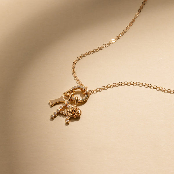 A gold chain necklace with a round ring in the center, holding a dog bone charm, an initial A charm with clear accent stones, and a flower charm.