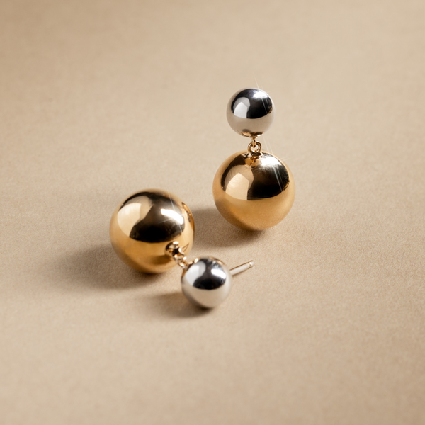 A pair of stud earrings with a silver round ball and a larger round gold ball dangling from it, set against a textured beige background.